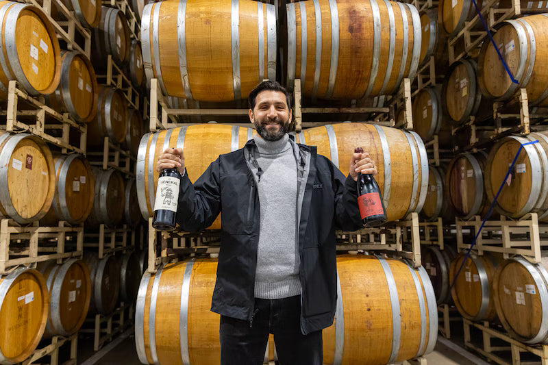 Diego Valeri holding two bottles of wine in a wine cellar with wooden barrels.