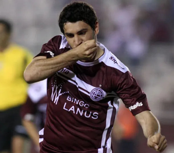 Soccer player in maroon and white jersey with 'Lanus' on a blurred stadium background