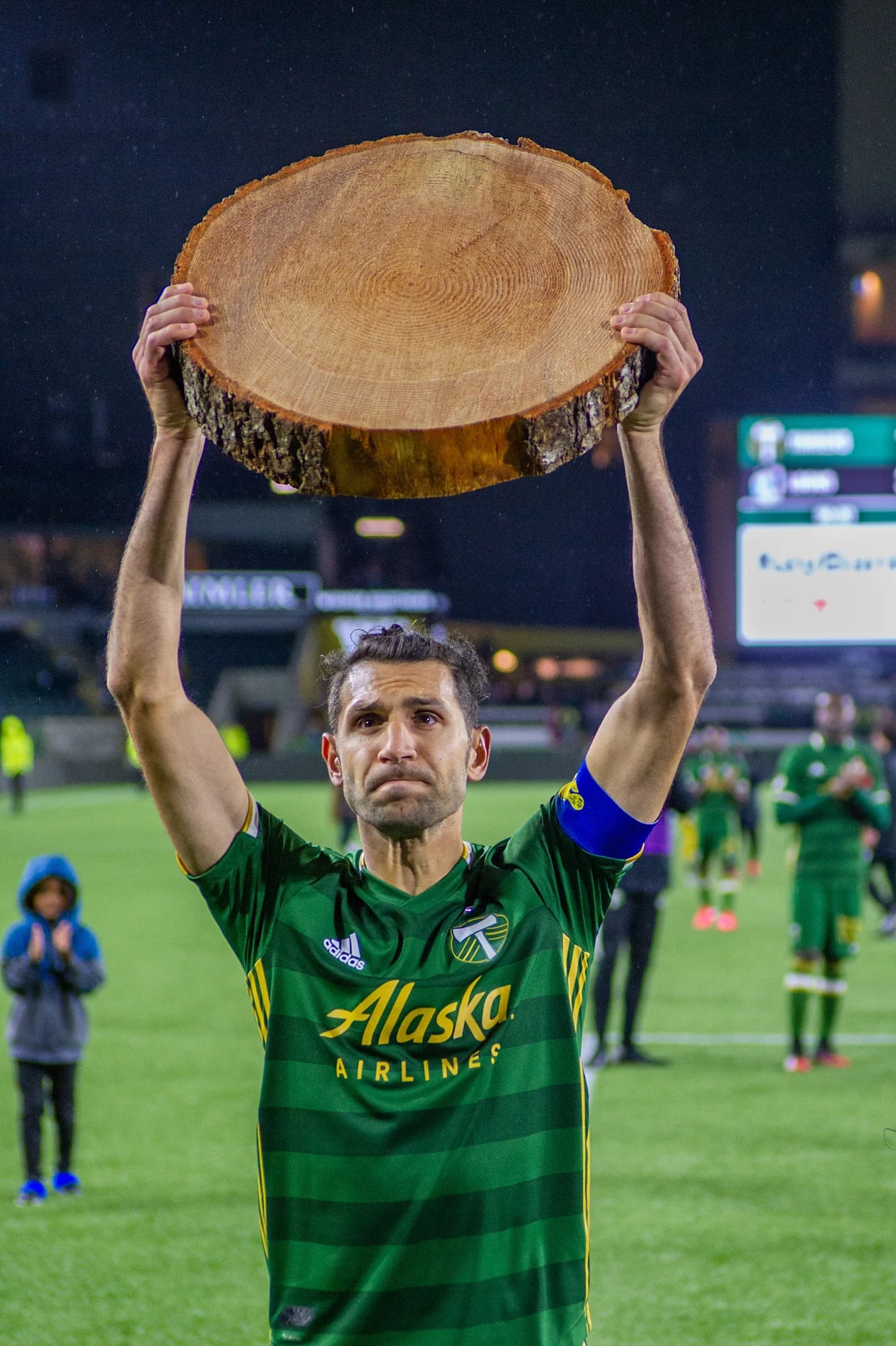 Person in green sports jersey holding a large wooden log aloft on a sports field. Portland Timbers - Diego Valeri