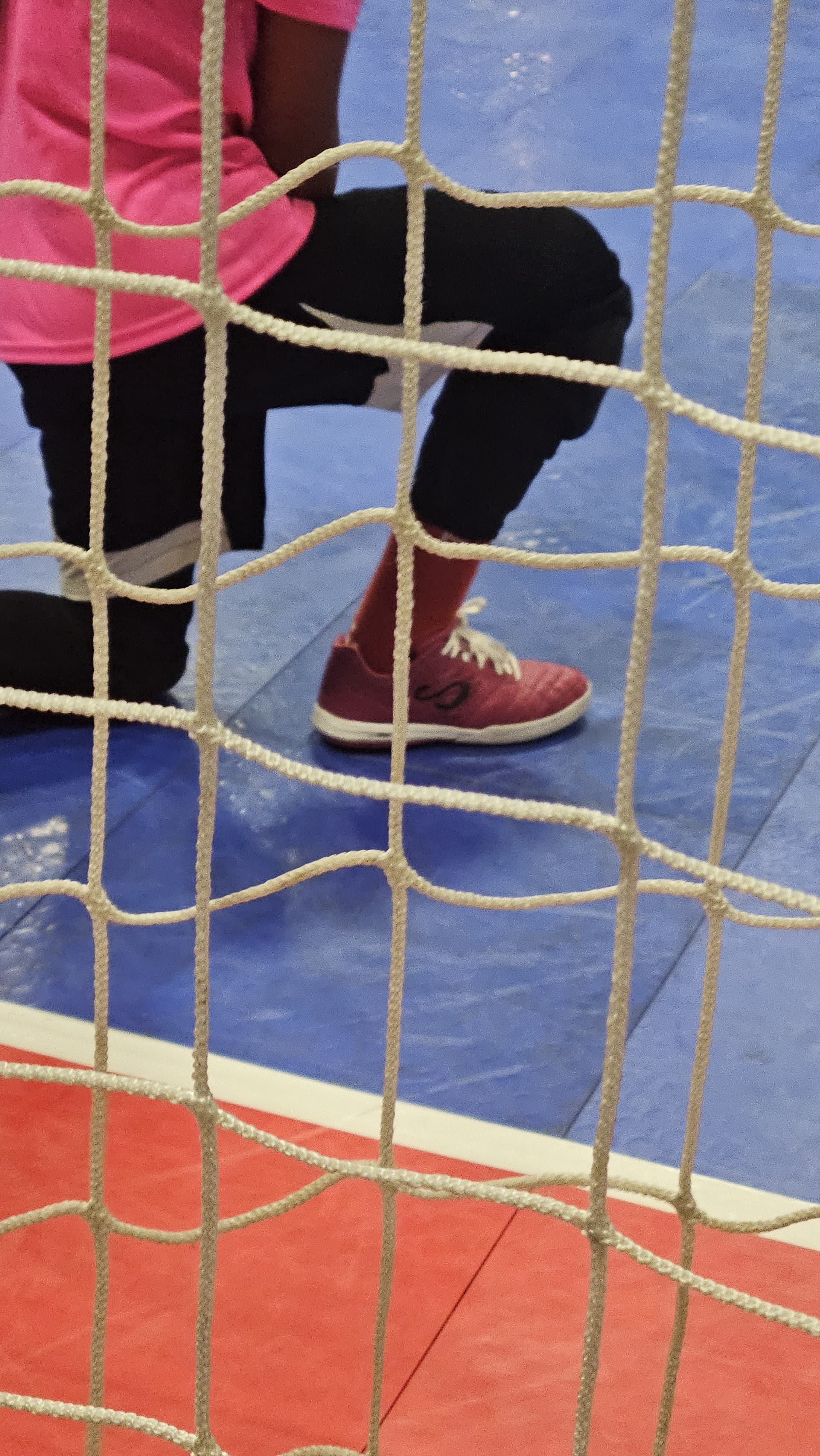 Goalkeeper wearing futsal purple shoes and black pants on a blue court, viewed through a white rope net.