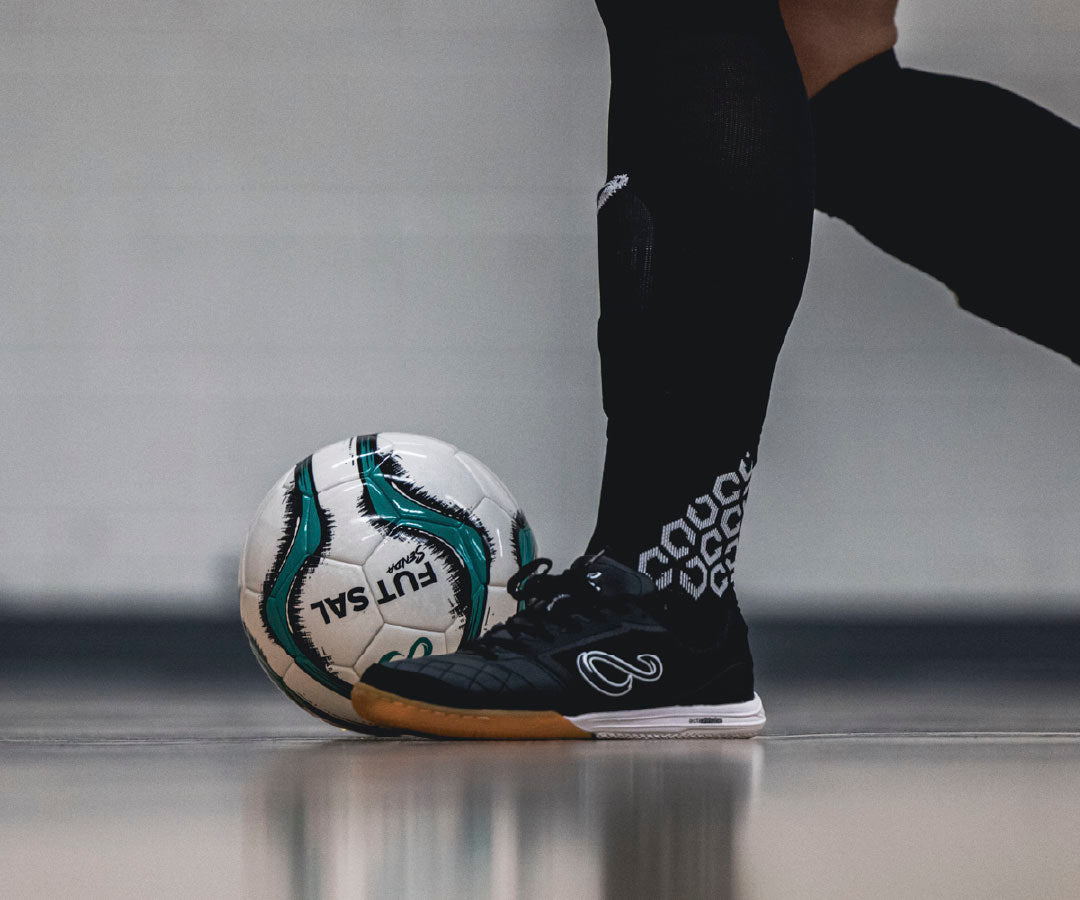Person wearing black futsal shoes with white patterns, kicking a white professional futsal ball on a gray surface.