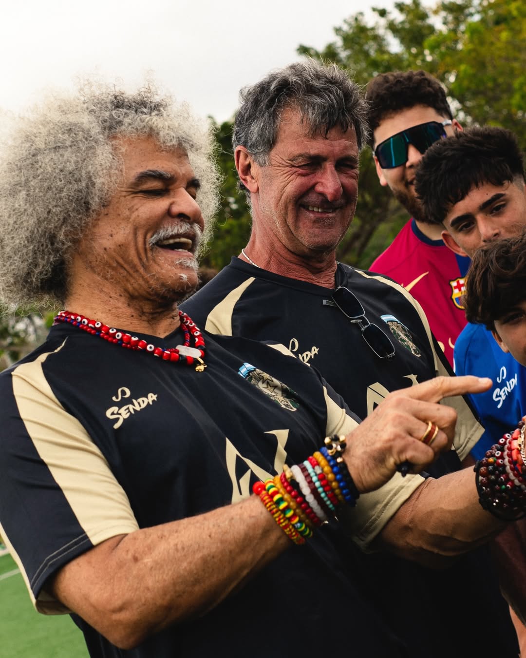 Mario Kempes and Carlos Valderrama on a sports field, one pointing towards a microphone held by another person; both with Senda gear.