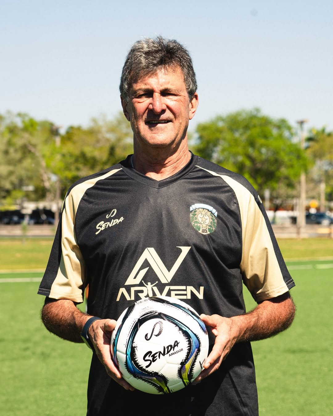 Mario Kempes holding a Senda soccer ball on a sports field with trees in the background; at Mario Kempes Soccer Clinic in Miami.
