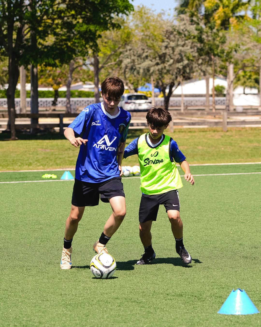 Two children playing soccer on a grass field with trees and buildings in the background. Senda gear at Mario Kempes Soccer Clinic.