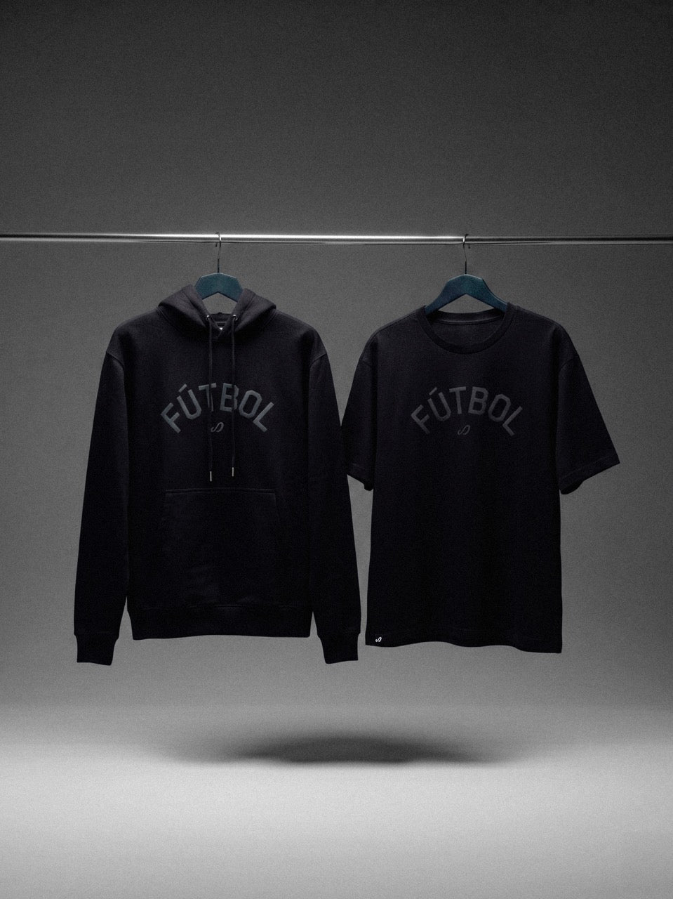 Black hoodie and t-shirt with 'FÚTBOL' text on hangers against a gray background. Senda Lifestyle Apparel Collection