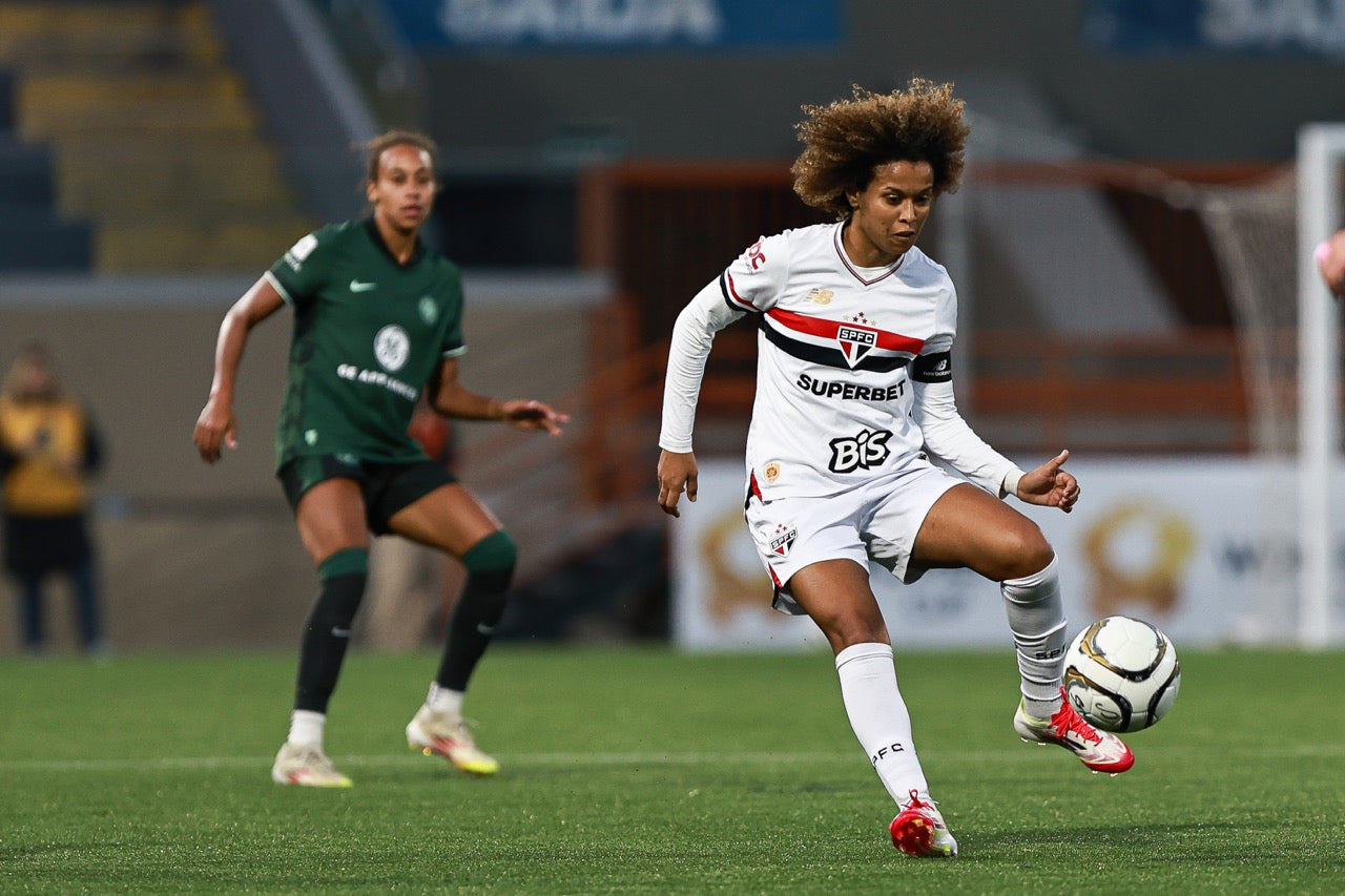 Two female soccer players in action on a field with one player in a white uniform kicking a soccer ball.