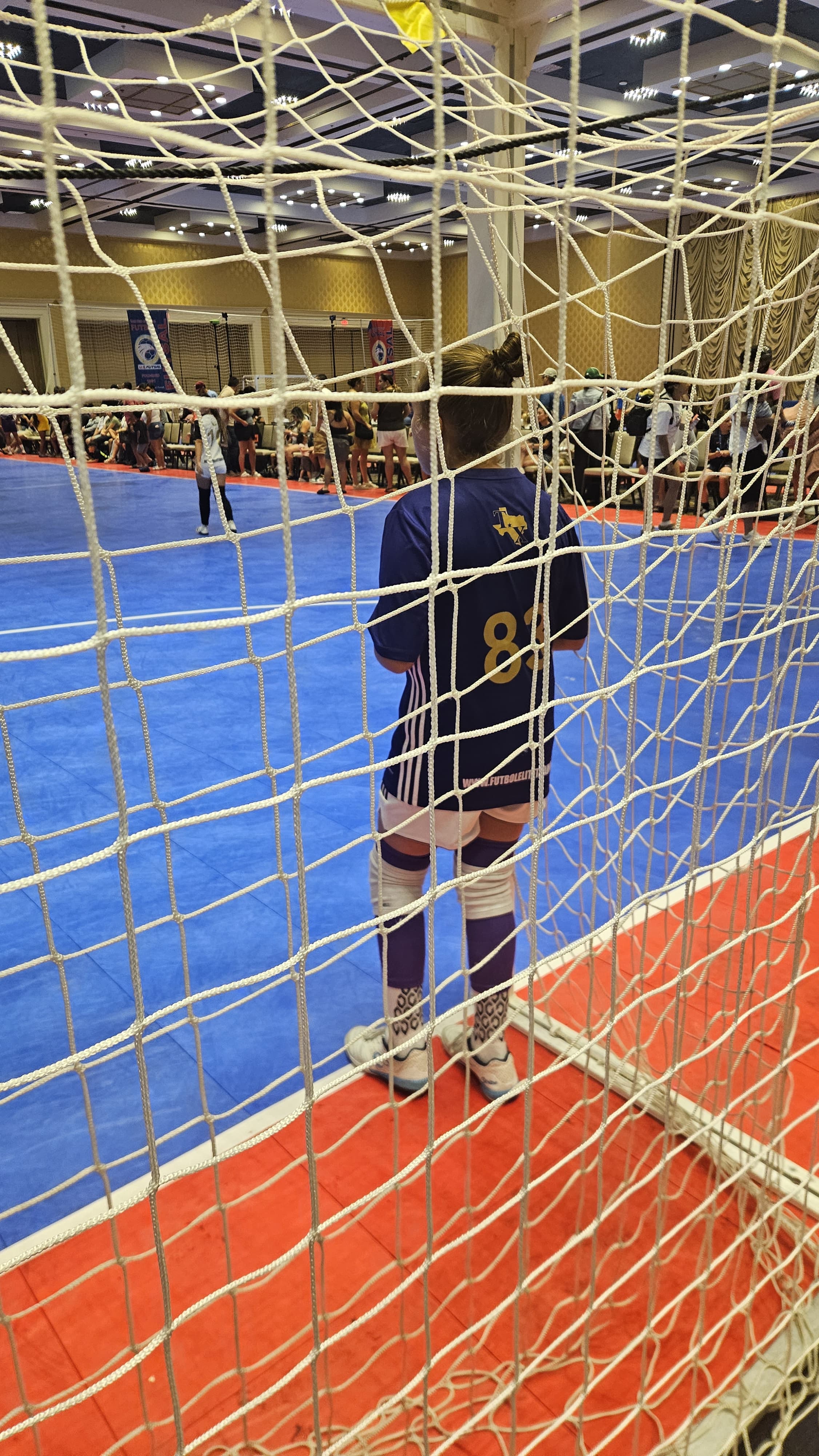 Goalkeeper  in a blue sports jersey standing behind a net on a blue and orange futsal court.