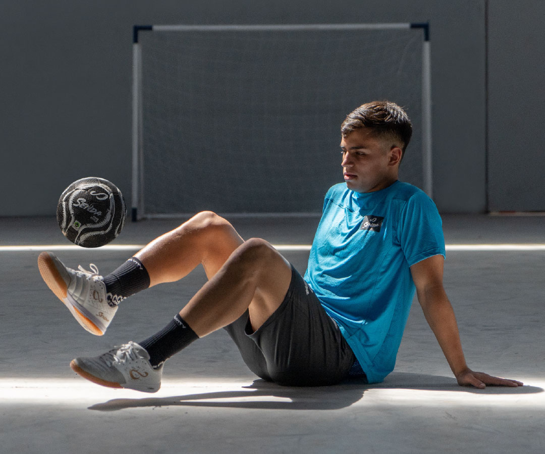 Person sitting on a concrete surface with a soccer ball, wearing a blue shirt and black shorts.
