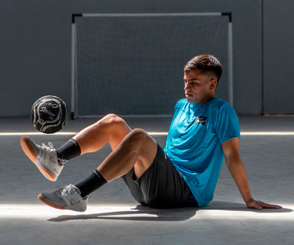 Person sitting on a concrete surface with a soccer ball, wearing a blue shirt and black shorts.