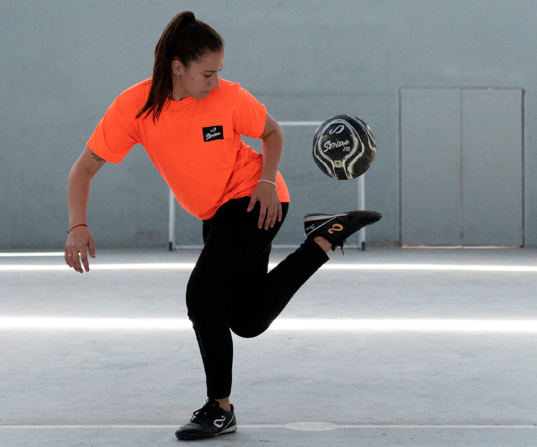 Person in orange shirt and black pants performing a soccer trick with a ball on a gray floor.
