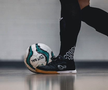 Person wearing black futsal shoes with white patterns, kicking a white professional futsal ball on a gray surface.