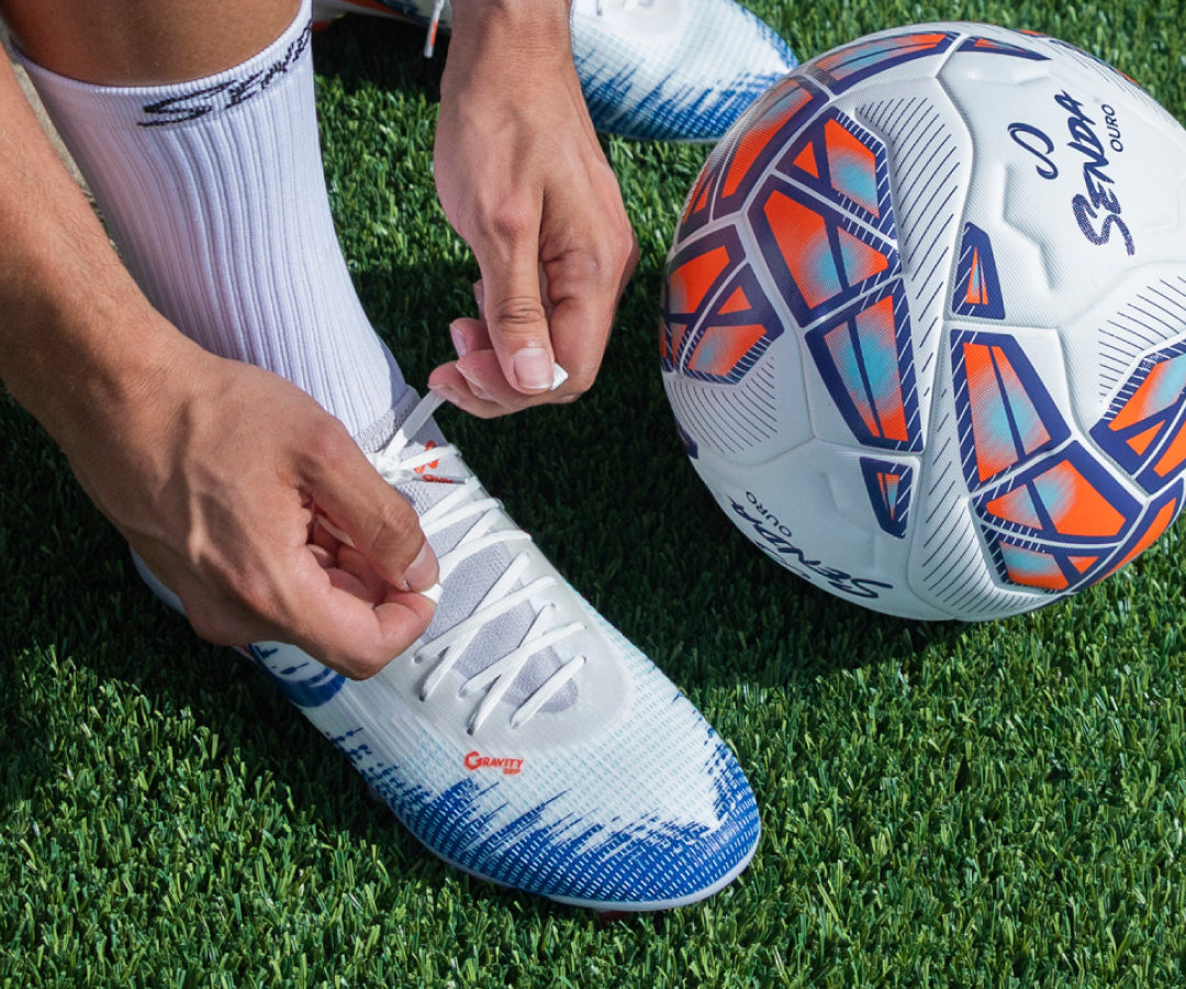 Person tying a Senda soccer cleat on grass with a soccer ball nearby