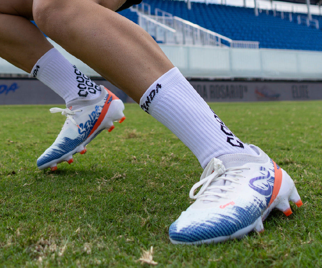 Person wearing white Senda soccer cleats with blue and orange accents on a grass field.