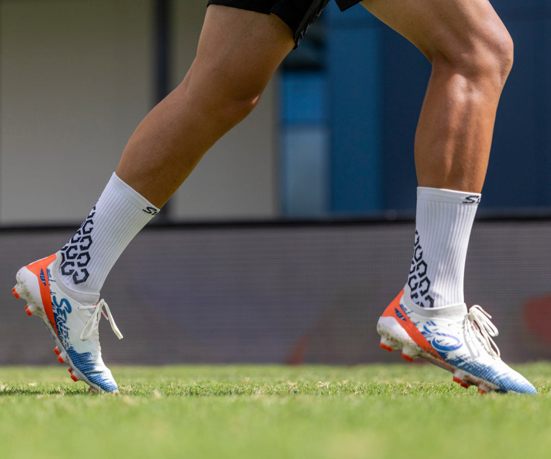 Close-up of soccer player's feet wearing orange and blue Senda cleats on a grass field.